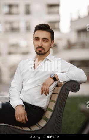 Bell'uomo d'affari seduto sulla panchina all'aperto durante il pranzo. Un giovane in una camicia bianca e con un orologio alla moda sulla mano che si pone all'aperto Foto Stock