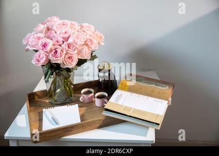 Rosa Bianco Rosa o'hara. Bouquet di rose rosa in vaso di vetro con una teiera bianca e due tazze di cappuccino su un vassoio su un tavolino tra i libri. Foto Stock