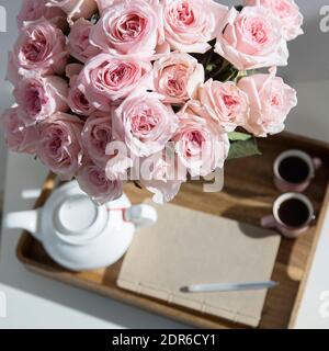 Rosa Bianco Rosa o'hara. Bouquet di rose rosa in vaso di vetro con una teiera bianca e due tazze di cappuccino su un vassoio su un tavolino tra i libri. Foto Stock