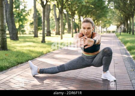 Preparazione. Foto a tutta lunghezza di attraente donna sportiva in abbigliamento sportivo che fa esercizio caldo mentre si allenano in un parco verde in un'estate soleggiata Foto Stock