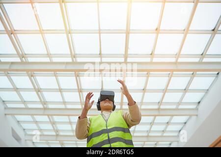 Ritratto grafico a basso angolo di un ingegnere femminile che indossa un visore VR mentre visualizza il progetto di progettazione architettonica in piedi sotto il tetto di vetro, spazio di copia Foto Stock