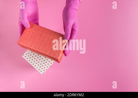 Mani femminili in guanti medici che tengono la scatola di regalo su sfondo rosa. Vista dall'alto. Primo piano. Spazio di copia. Concetto di sicurezza gifting. Foto Stock