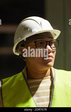 Ombra e ritratto chiaro di una moderna ingegnere femminile che indossa un cappello e guardando via mentre ispezionano il luogo di costruzione Foto Stock