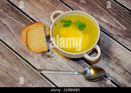 Un piatto profondo di brodo di pollo con mezzo uovo e un rametto di prezzemolo si trova su un tavolo di legno, accanto ad un cucchiaio e due fette di pane bianco. Piatto la Foto Stock