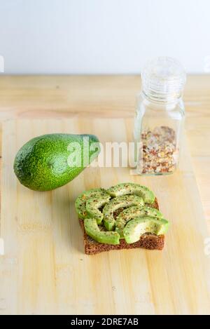 Toast piccante all'avocado - colazione sana. Pane tostato alla segale con avocado verde a fette, succo di lime, sale, spezie - miscela di pepe-mais su un asse di legno. Vista dall'alto. Foto Stock