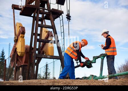 Vista completa di due operatori petroliferi che indossano tute, giubbotti arancioni e caschi, che lavorano sul campo dell'olio accanto al martinetto della pompa dell'olio, utilizzando una chiave industriale, il bullone di serraggio sul tubo, l'altro facendo delle note Foto Stock