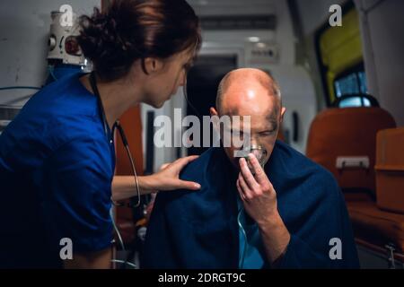 Un uomo ferito che preme una maschera di ossigeno al viso, una paramedica femminile sta cercando di calmarlo. Foto Stock