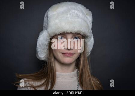 Bella ragazza bionda giovane con un cappello invernale in pelliccia bianca all'interno su uno sfondo nero, primo piano ritratto Foto Stock