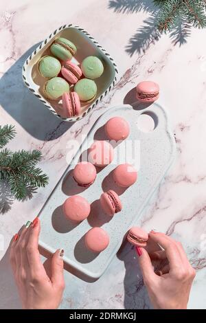 Macaroni rosa e verde menta su un vassoio e in una ciotola di ceramica. Le mani femminili tengono uno spuntino gustoso. Gustoso dessert creativo in verde e rosso su tavolo di marmo wi Foto Stock