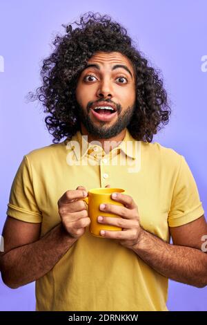 emozionale arabo maschio con tazza in mani, isolato su sfondo viola. bel ragazzo con capelli ricci guardare la macchina fotografica con gli occhi aperti, sorpreso Foto Stock