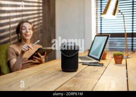 Donna che parla con l'altoparlante intelligente mentre lavora sul tablet digitale in un comodo ufficio domestico. Concetto di una casa intelligente e intelligenza artificiale Foto Stock