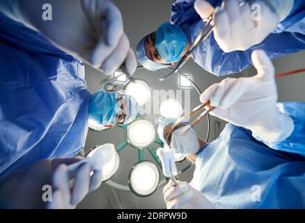 Vista dal basso dei medici che detengono strumenti medici durante la chirurgia plastica. Indossare maschere facciali di protezione e guanti sterili mentre si lavora in sala operatoria. Concetto di medicina. Foto Stock