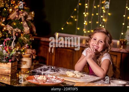 Una ragazza sorridente è seduta ad un tavolo nella cucina decorata, con pasta cruda sul tavolo davanti a lei. Il naso è cosparso di farina. Selettivo Foto Stock