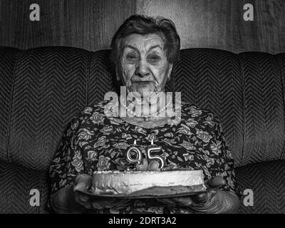 Un colpo in scala di grigi di una donna di 95 anni che tiene il suo compleanno torta mentre si siede sul divano all'interno Foto Stock