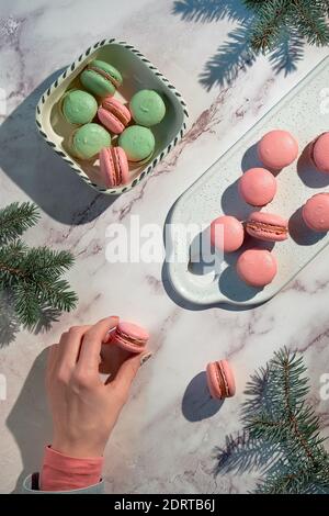 Macaroni rosa e verde menta su un vassoio e in una ciotola di ceramica. Gustoso dessert creativo in verde e rosso su tavolo di marmo con ramoscelli di abete femminile e invernale. Foto Stock