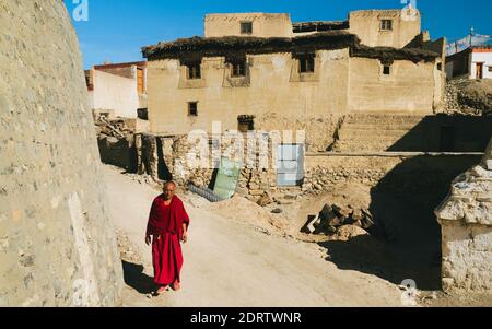 Monaco buddista in abiti rossi cammina lungo la strada sterrata fiancheggiata da case di mattoni di fango con tetti di paglia nel villaggio di Kibber, Himachal Pradesh, India. Foto Stock