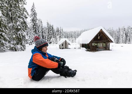 Ragazzo seduto sulla neve di fronte a una tradizionale capanna di legno in un prato alpino coperto di neve nelle alpi Giulie in inverno, Pokljuka, Slovenia Foto Stock