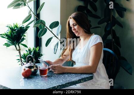 Una donna incinta affascinante sta leggendo un e-libro ad un tavolo in un caffè. Foto Stock