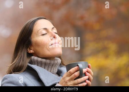 Donna adulta rilassata che tiene la tazza di caffè respirando aria fresca dentro autunno in una foresta Foto Stock