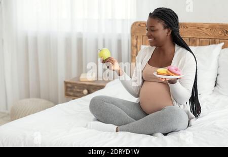 Nutrizione prenatale. Donna incinta nera scelta tra mela e ciambelle a casa Foto Stock