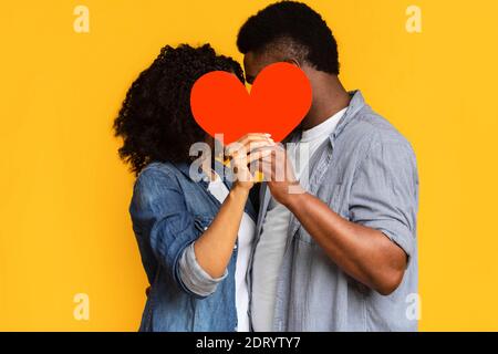 Amore segreto. Romantica coppia nera nascondendosi e baciandosi dietro cuore di carta rossa Foto Stock