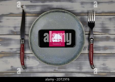 Viersen, Germania - 9 maggio. 2020: Primo piano dello schermo del telefono cellulare con la scritta del logo del servizio di consegna del cibo foodora su tavolo di legno con piatto e cutle Foto Stock