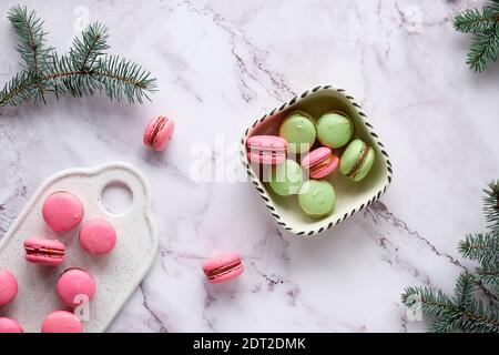 Macaroni rosa e verde menta su un vassoio e in una ciotola di ceramica. Gustoso dessert creativo in verde e rosso su tavolo in marmo con rametti di abete. Foto Stock