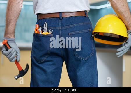 Primo piano di vari attrezzi manuali idraulici nella tasca posteriore dei jeans dell'uomo. Riparatore professionista che tiene in mano un martello e un casco Foto Stock