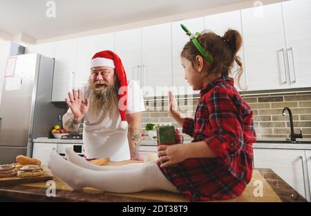 Un seduto su un tavolo figlia e padre nel cappello di Babbo Natale e armi tatuate agitando l'uno verso l'altro. Concetto di relazione familiare Foto Stock