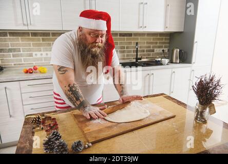 Divertente uomo bearded che indossa la cucina del cappello di Santa in cucina a casa. Buon Natale e Felice Anno Nuovo. Cucina e concetto di vacanza Foto Stock