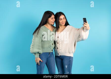 Le sorelle gemelle sorridono e prendono un selfie su un telefono cellulare. Sfondo blu e spazio laterale vuoto. Foto Stock