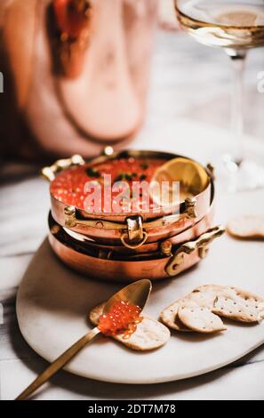 Caviale rosso in piatti di rame metallico con cracker salati e bicchiere di champagne a bordo su tavola di marmo bianco, fuoco selettivo. Concetto di umore per le vacanze invernali Foto Stock