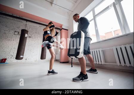 Una giovane ragazza di kickboxer forte calcia il suo partner scintillante con una zampa di pugilato in una sessione di allenamento personale Foto Stock