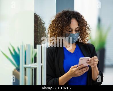 Donna d'affari che indossa la maschera facciale utilizzando lo smartphone in ufficio Foto Stock