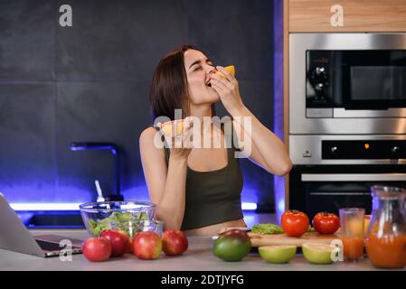 Ragazza piuttosto snella che mangia pompelmo amaro o arancia mentre prepara una deliziosa e sana colazione alla frutta in cucina contemporanea. Foto Stock