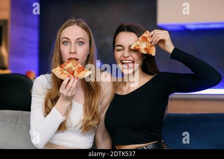 Gioiose giovani donne in soggiorno giocando con fette di pizza, sorridendo e coprendo gli occhi. Consegna pizza online. Foto Stock