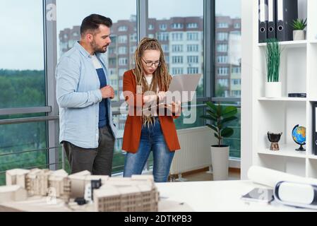 Piacevole esperienza moderna elegante e soddisfatto giovani designer discutere momenti di lavoro utilizzo di un progetto con progetto di nuovi edifici vicino a panoramic win Foto Stock