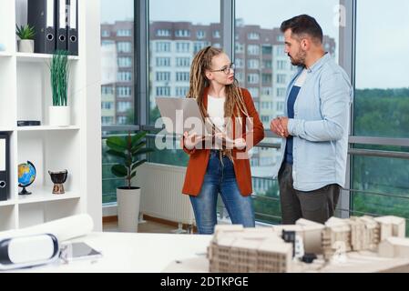 Piacevole esperienza moderna elegante e soddisfatto giovani designer discutere momenti di lavoro utilizzo di un progetto con progetto di nuovi edifici vicino a panoramic win Foto Stock