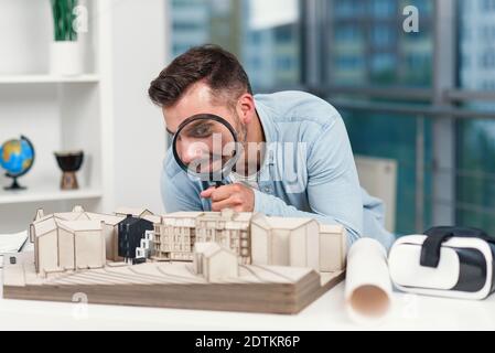 Divertente immagine di architetto guarda attraverso la lente di ingrandimento a un modello di casa. Ispezione della casa e concetto di bene immobile. Foto Stock