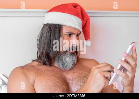 Vista ravvicinata di bearded con capelli lunghi uomo caucasico nel cappello di Babbo Natale usa un PC tablet che si posa sul letto. Concetto di vacanza, festa e nuovo anno. Foto Stock