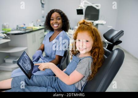 Bambino presso l'ufficio del dentista. Bambina paziente con capelli rossi, seduta in sedia odontoiatria, guardando la fotocamera e puntando su tablet con denti panoramici Foto Stock