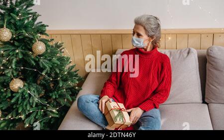 Donna anziana triste che indossa la maschera seduta sul divano da solo tenendo un regalo di natale . Stile di vita pandemico Covid-19 Foto Stock