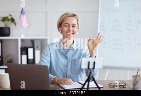 Positiva insegnante di sesso femminile che dà lezioni di inglese online su smartphone, mostrando un gesto OK a casa Foto Stock