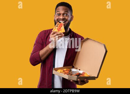 African Man mangiare Pizza sorridendo a Camera su sfondo giallo Foto Stock