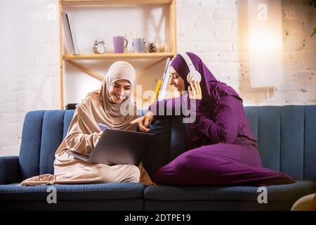 Laughing. Felice e giovane due donne musulmane a casa che parlano, sorridendo, divertendosi, seduti sul divano. Amicizia, tecnologia moderna, concetto di emozioni. Stile di vita e cultura, tradizioni arabe, etnia. Foto Stock