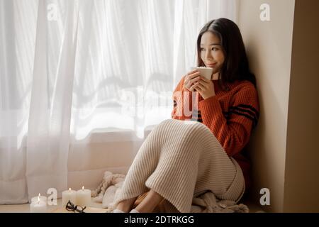 Giovane bella donna asiatica che tiene una tazza di caffè e marshmallows sulla parte superiore, seduto a casa e guardando fuori la finestra. Felice ragazza che beve cioccolato in maglione in inverno freddo Foto Stock