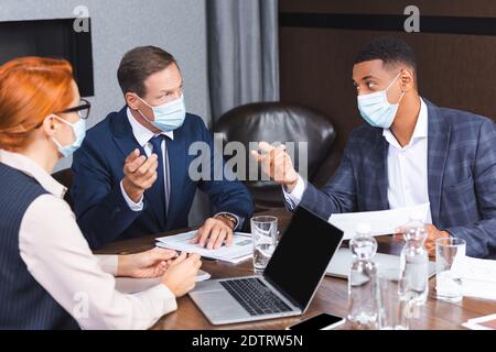 Uomini d'affari multiculturali in maschere mediche che gesturing durante la discussione vicino a una collega femminile sul posto di lavoro, banner Foto Stock