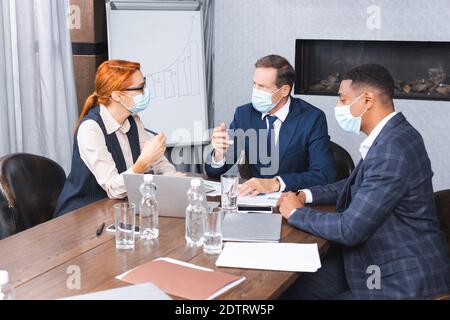 Uomini d'affari multiculturali in maschere mediche che parlano con l'investitore mentre si siede sul luogo di lavoro con dispositivi digitali Foto Stock