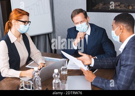 Uomini d'affari multiculturali in maschere mediche che parlano mentre si siedono sul posto di lavoro con dispositivi e documenti digitali Foto Stock