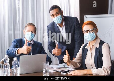 Uomini d'affari multiculturali in maschere mediche guardando la macchina fotografica mentre mostra pollice in su sul posto di lavoro Foto Stock